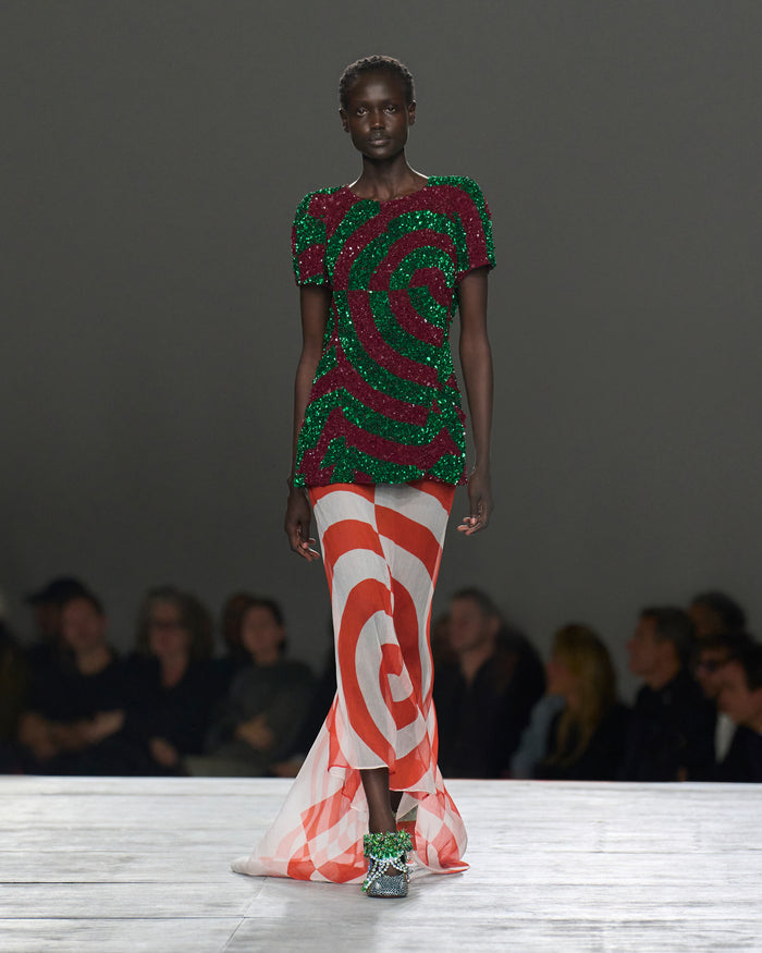 Model wearing a vibrant, patterned outfit on a runway with an audience in the background.