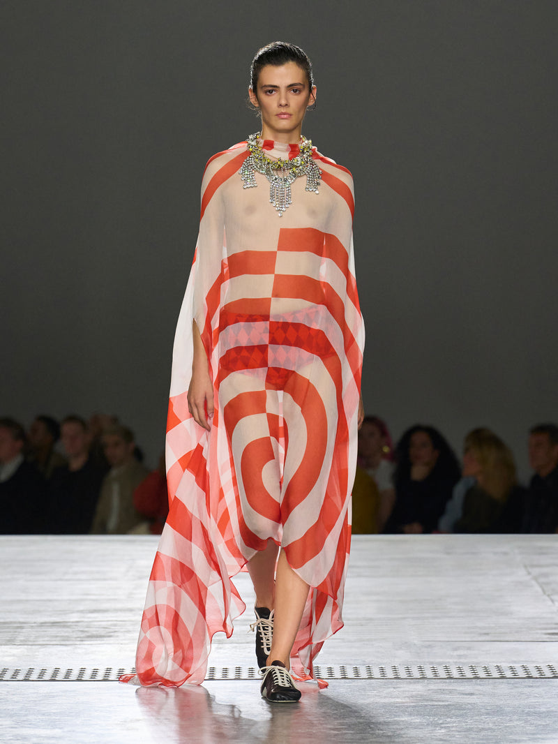 Model walking on a runway wearing a red and white striped dress with a blurred audience in the background.