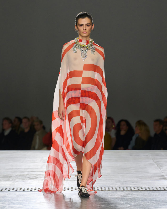 Model walking on a runway wearing a red and white striped dress with a blurred audience in the background.
