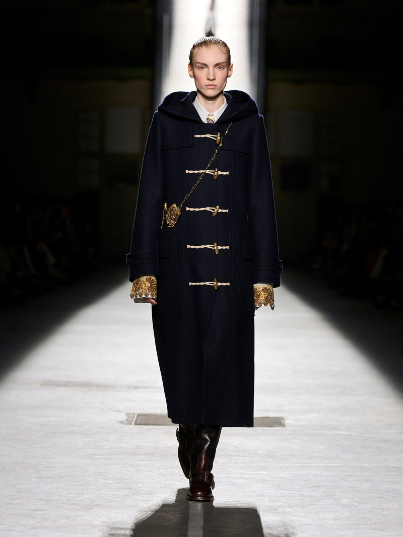 Model walking on a runway wearing a dark coat with gold buttons.