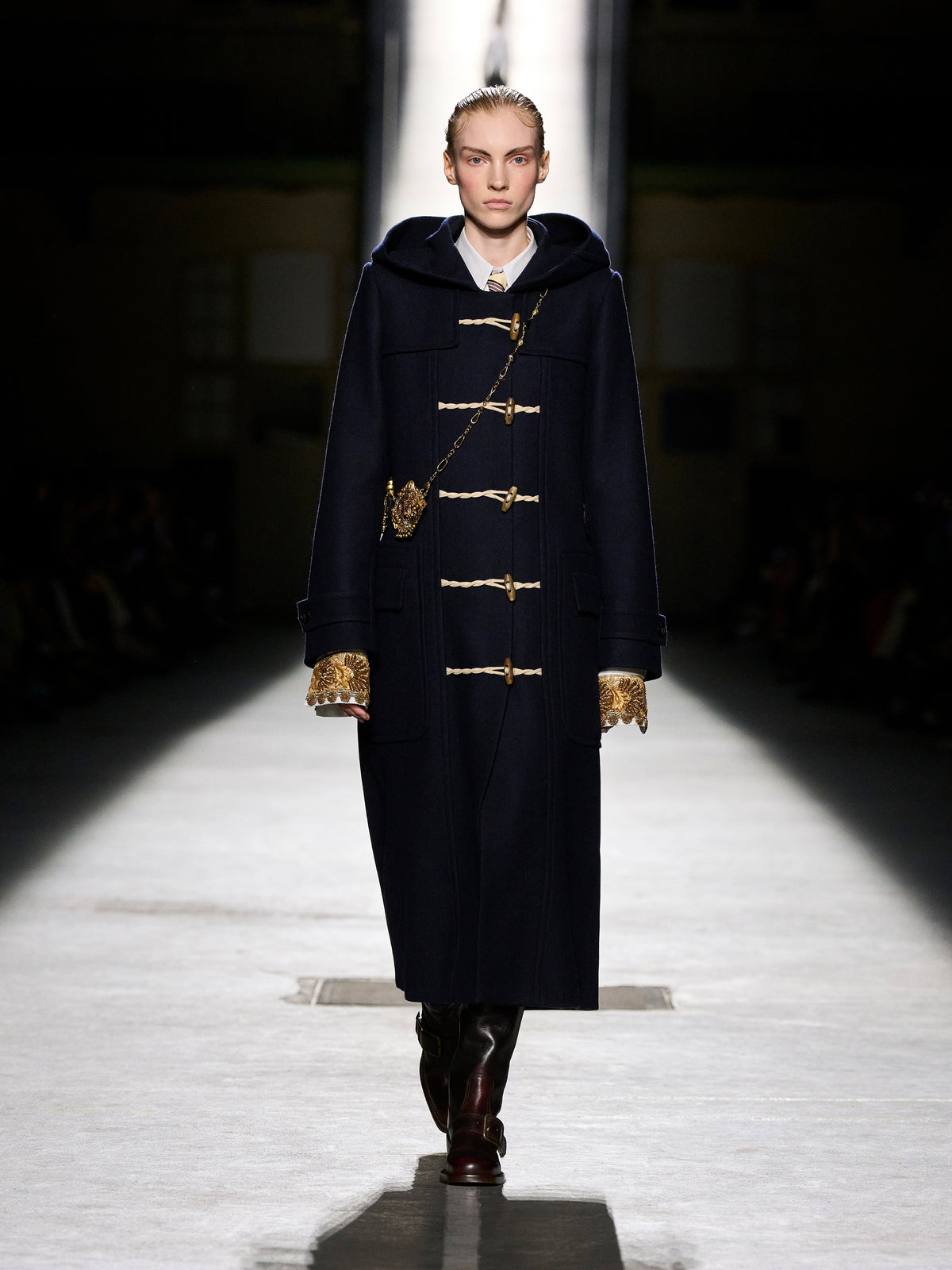 Model walking on a runway wearing a dark coat with gold buttons.