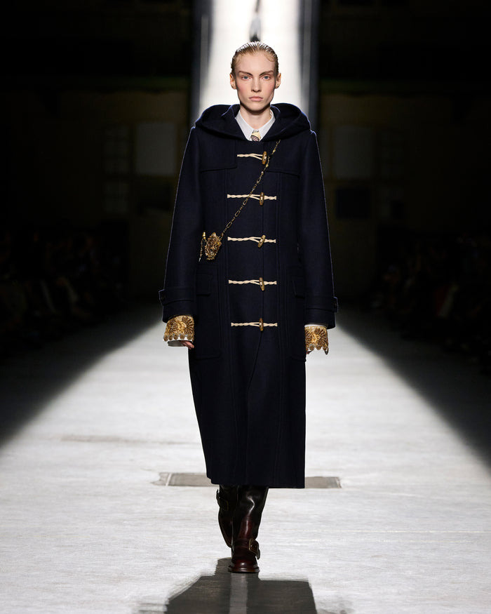 Model walking on a runway wearing a dark coat with gold buttons.