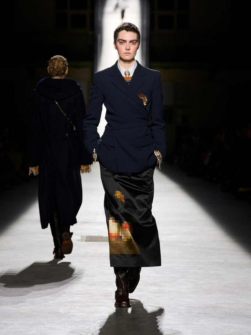 Model walking on a runway wearing a dark blue suit with gold accents.