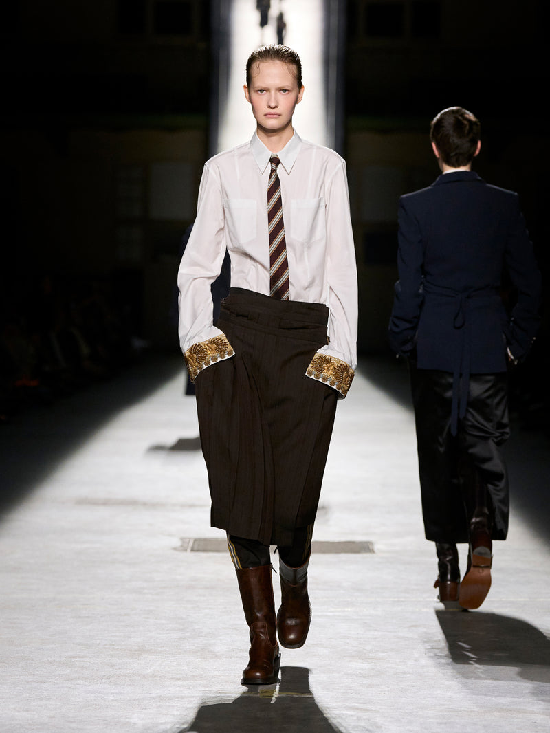 Model walking on a runway wearing a white shirt, brown tie, and dark coat with gold trim.