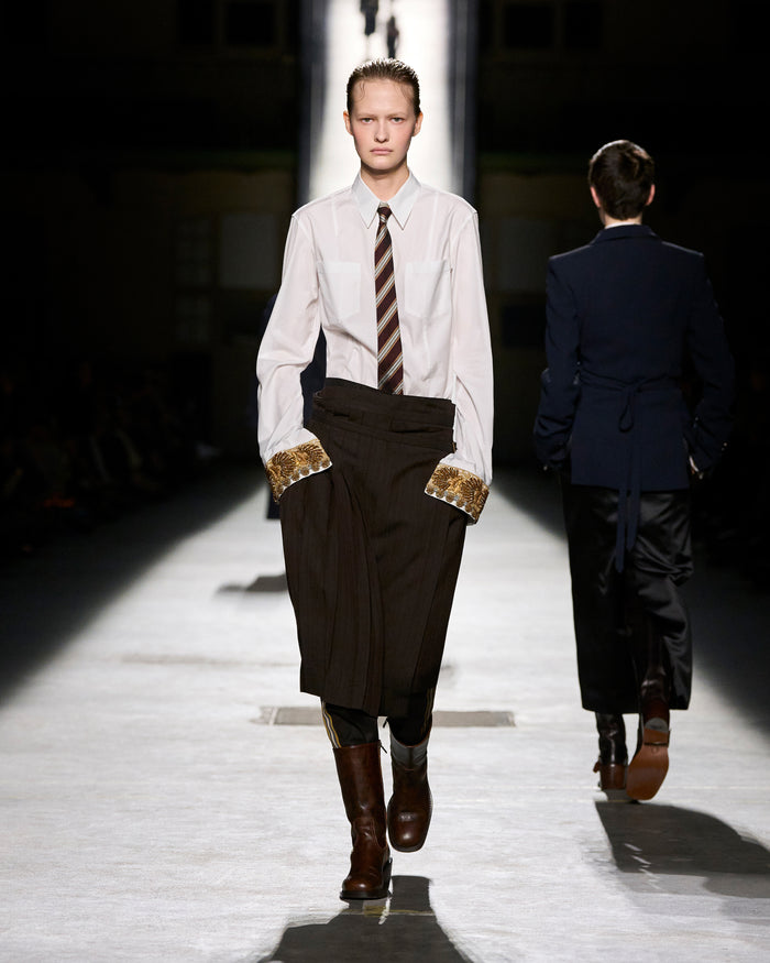 Model walking on a runway wearing a white shirt, brown tie, and dark coat with gold trim.