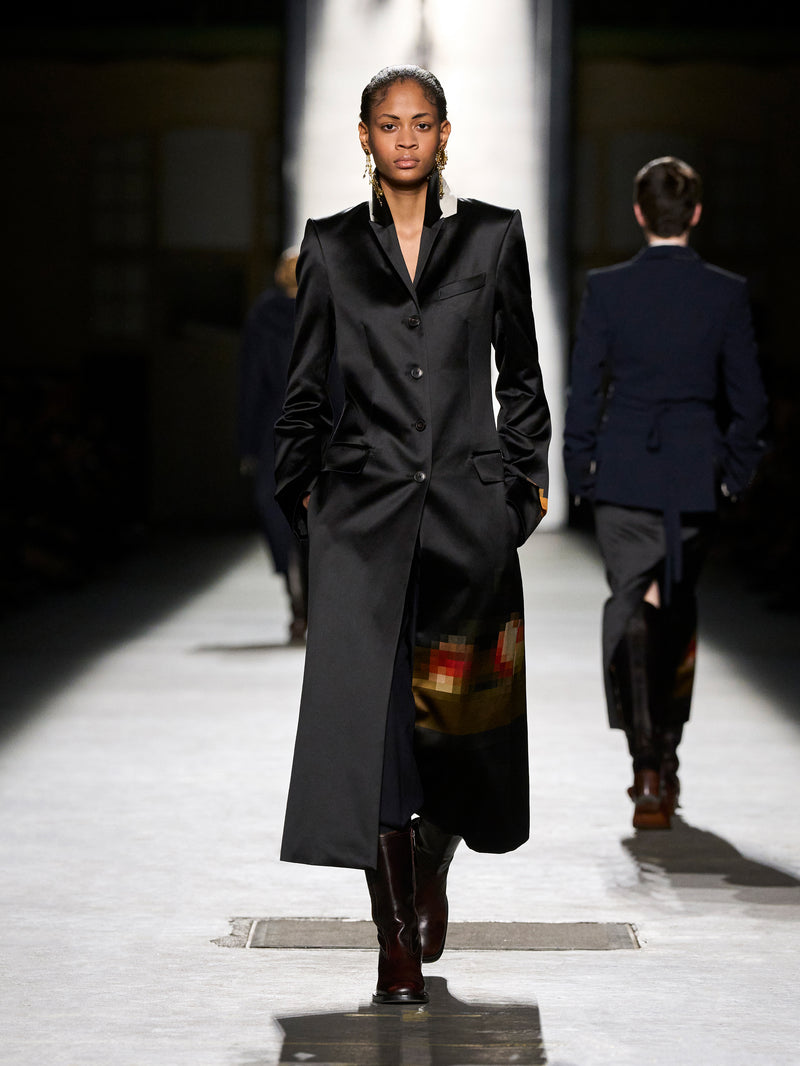 Model walking on a runway wearing a long black coat.