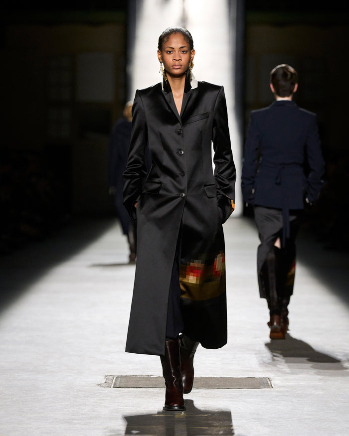 Model walking on a runway wearing a long black coat.