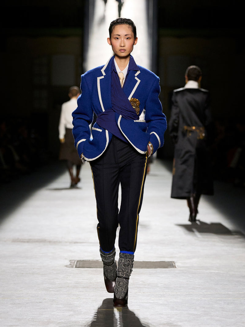 Model walking on a runway wearing a blue blazer with white trim and black pants.