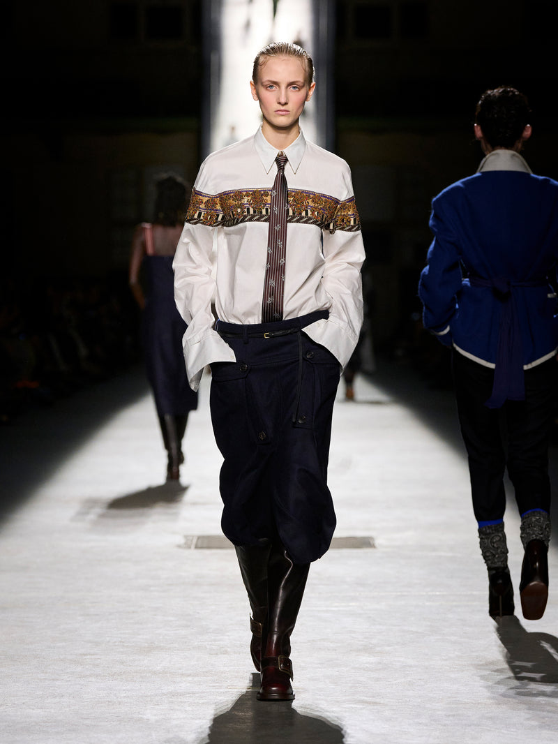 Model walking on a runway wearing a white shirt with a brown belt and dark pants.