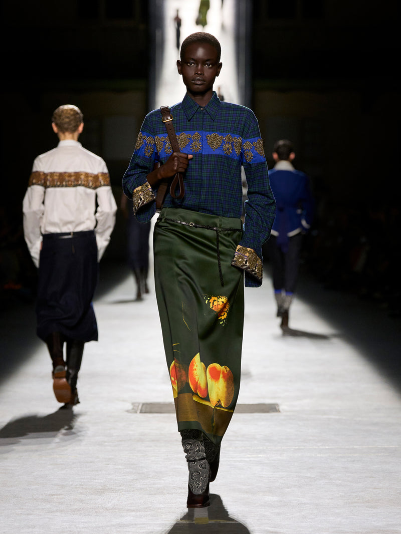 Model walking on a runway wearing a blue patterned sweater and green pants with fruit design.