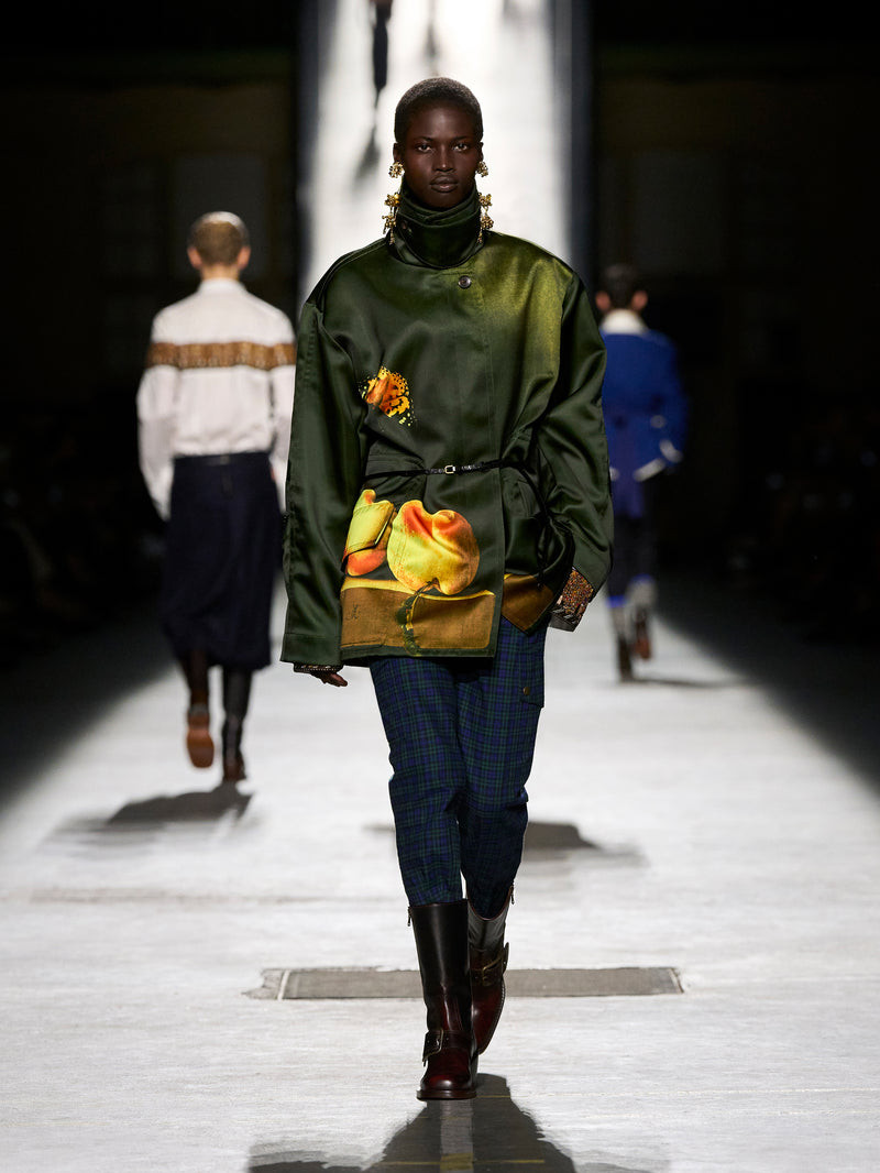 Model walking on a runway wearing a green jacket with floral design and blue jeans.