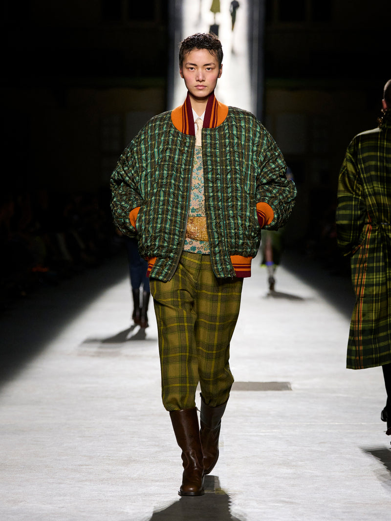 Model walking on a runway wearing a green plaid outfit with orange accents.