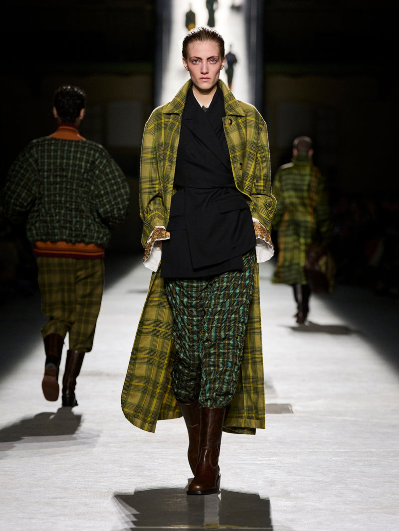 Model walking on a runway wearing a green plaid coat and skirt during a fashion show.