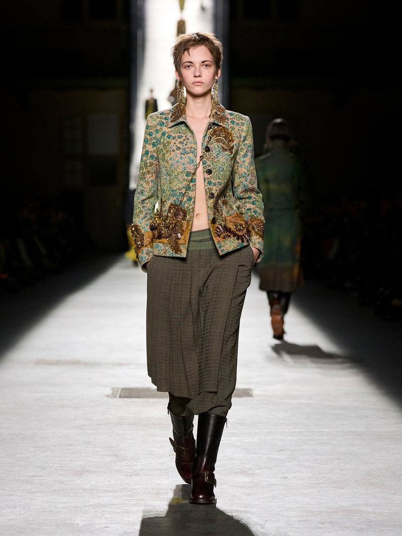 Model walking on a runway wearing a patterned jacket and plaid pants.
