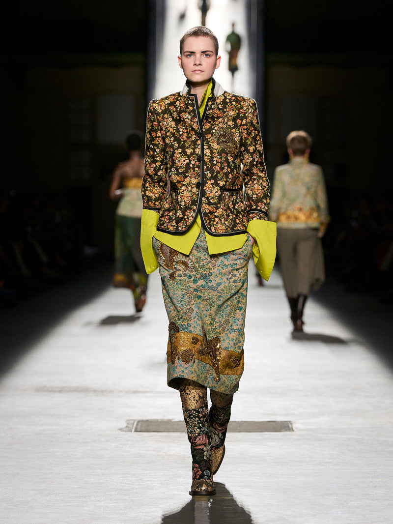 Model walking on a runway wearing a patterned outfit with a bright yellow jacket.