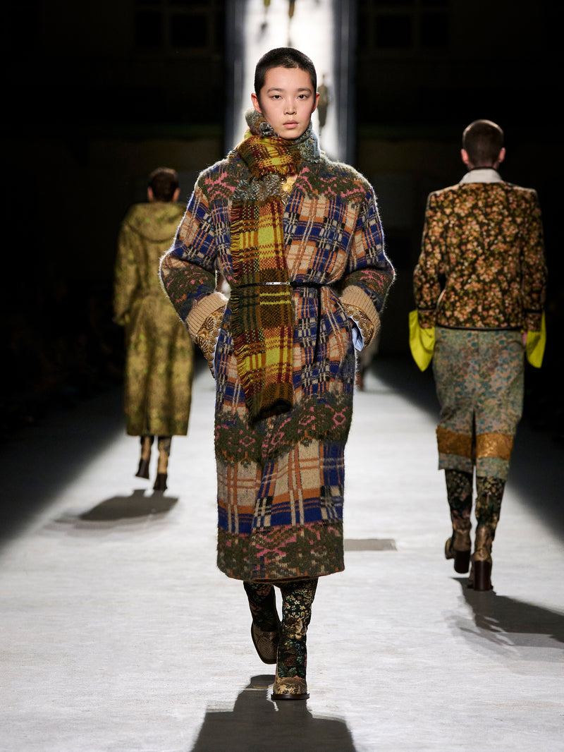 Model walking on a runway wearing a patterned coat with other models in the background.