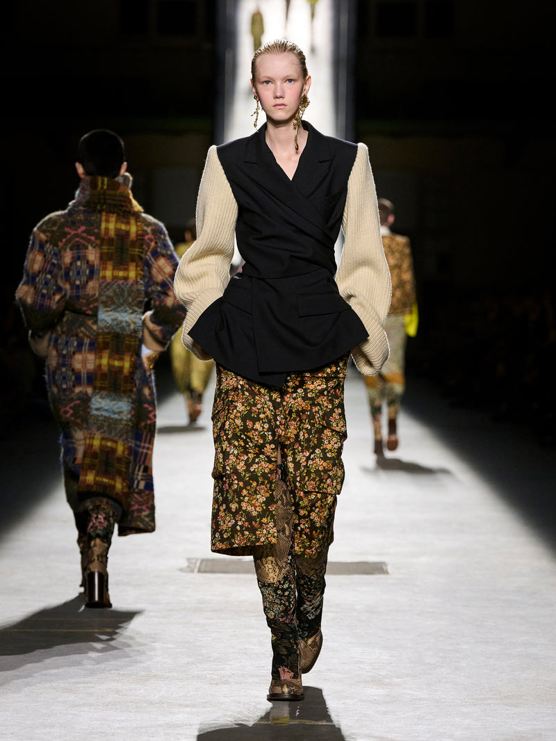 Model walking on a runway wearing a black and beige outfit with floral pants.