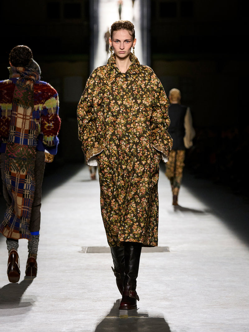 Model walking on a runway wearing a floral coat and patterned pants.