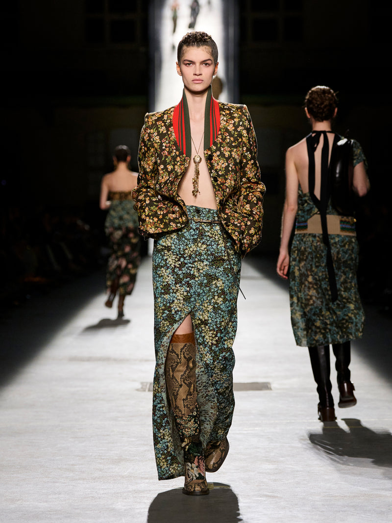 Model walking on a runway wearing a floral and snake print outfit with a red and green ribbon.