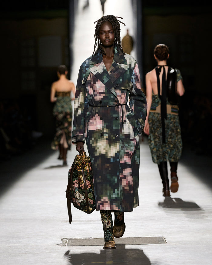 Model walking on a runway wearing a colorful, patterned outfit with a handbag.