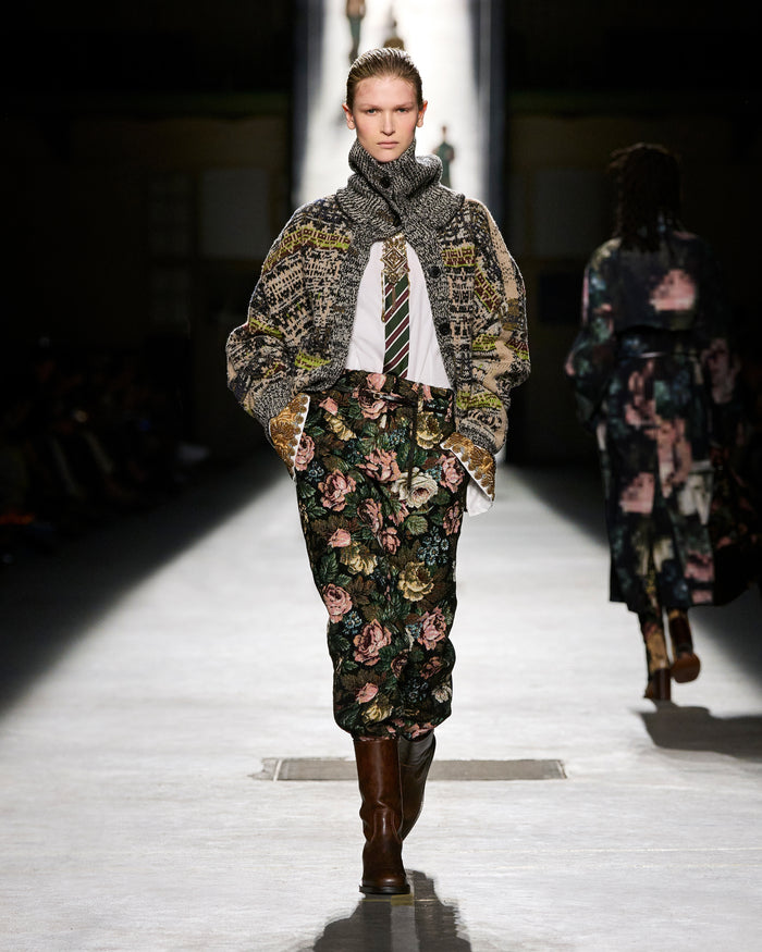 Model walking on a runway wearing a floral dress and patterned coat.