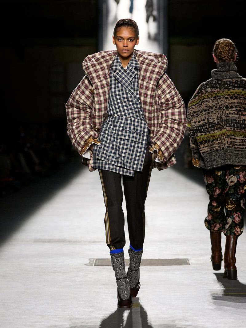 Model walking on a runway wearing a plaid coat and checkered dress with another model in the background.