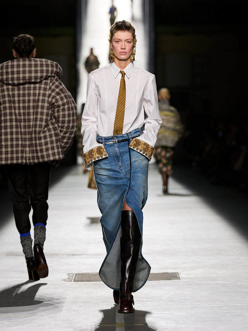 Model walking on a runway wearing a white shirt, blue jeans, and black boots.