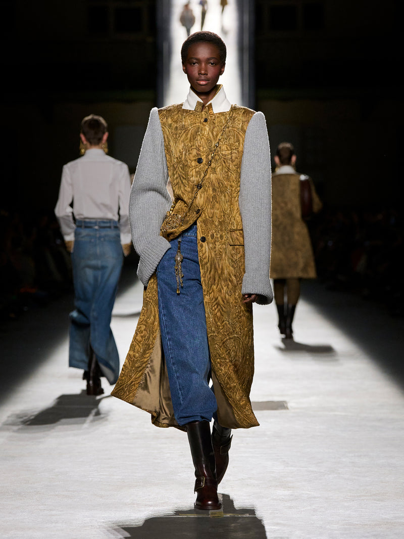 Model walking on a runway wearing a mustard yellow vest over a gray sweater and blue jeans.