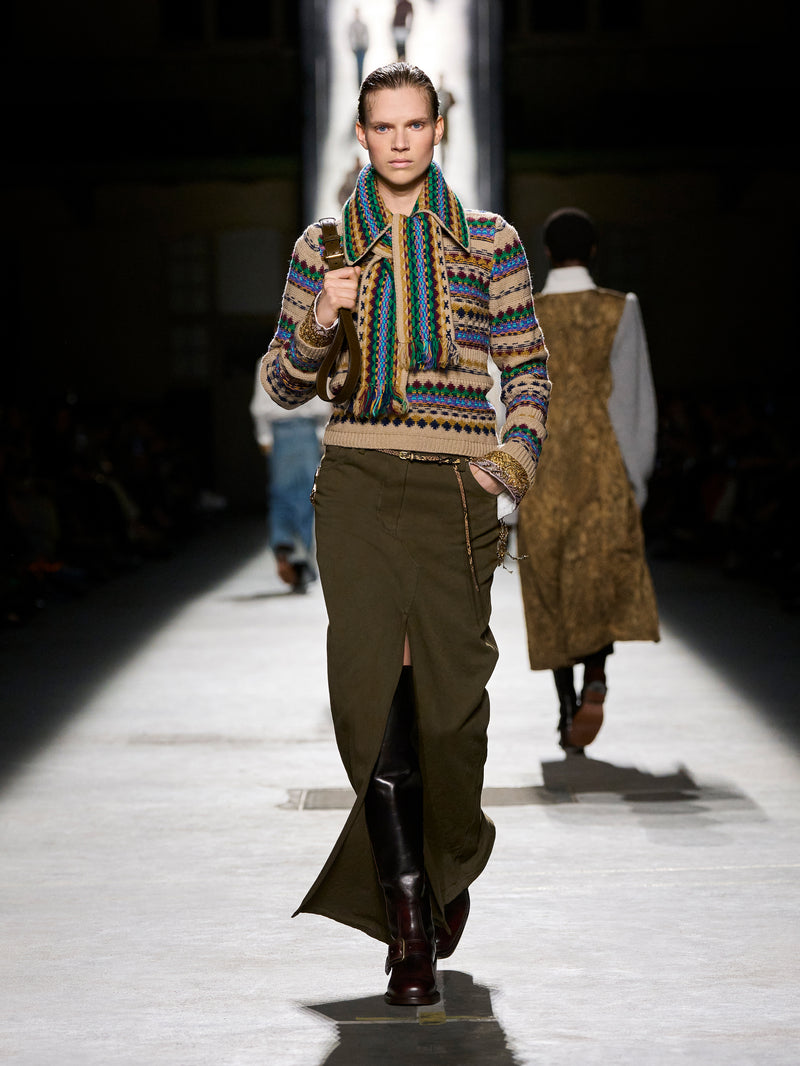 Model walking on a runway wearing a patterned sweater and green skirt.