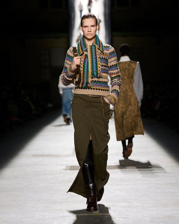 Model walking on a runway wearing a patterned sweater and green skirt.