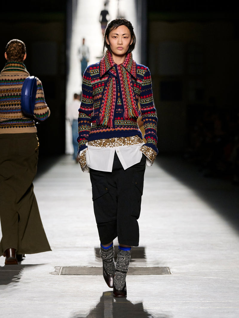 Model walking on a runway wearing a colorful patterned sweater and black pants.