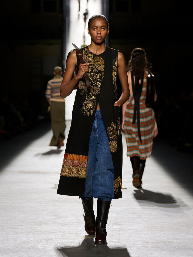 Model walking on a runway wearing a long, dark dress with floral patterns and a blue skirt.