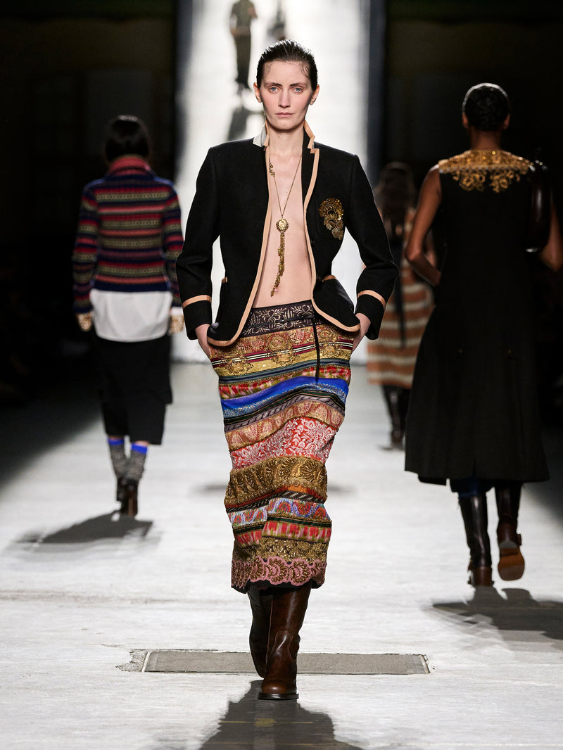 Model walking on a runway wearing a colorful dress with a black jacket.