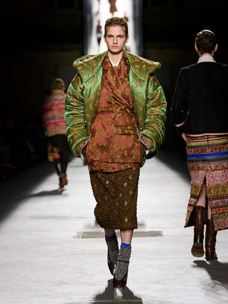 Model walking on a runway wearing a patterned outfit with a green jacket.