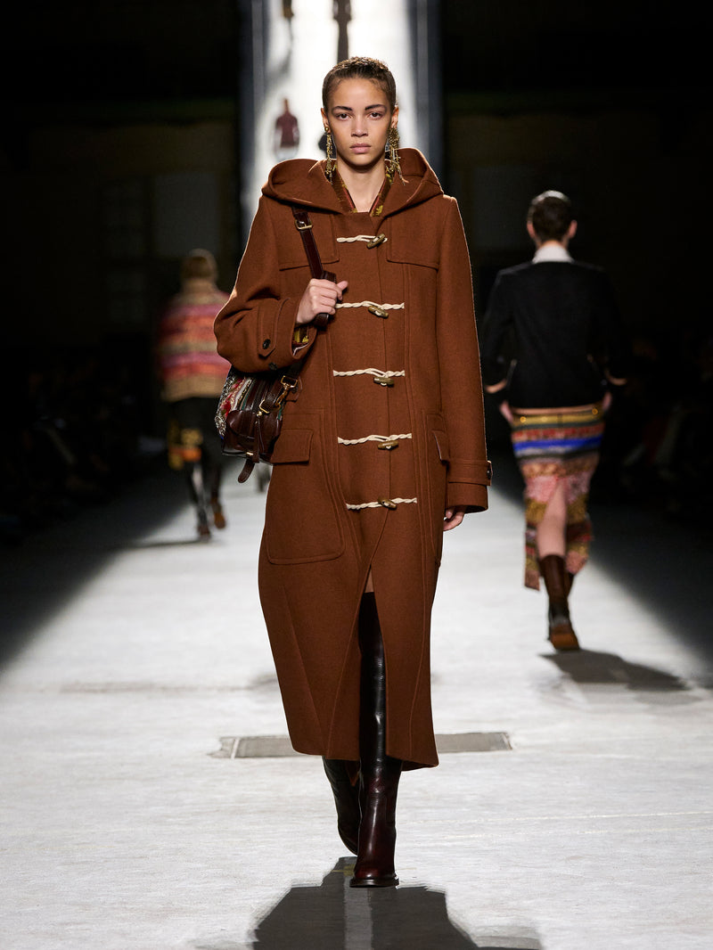 Model walking on a runway wearing a brown coat with gold buttons.
