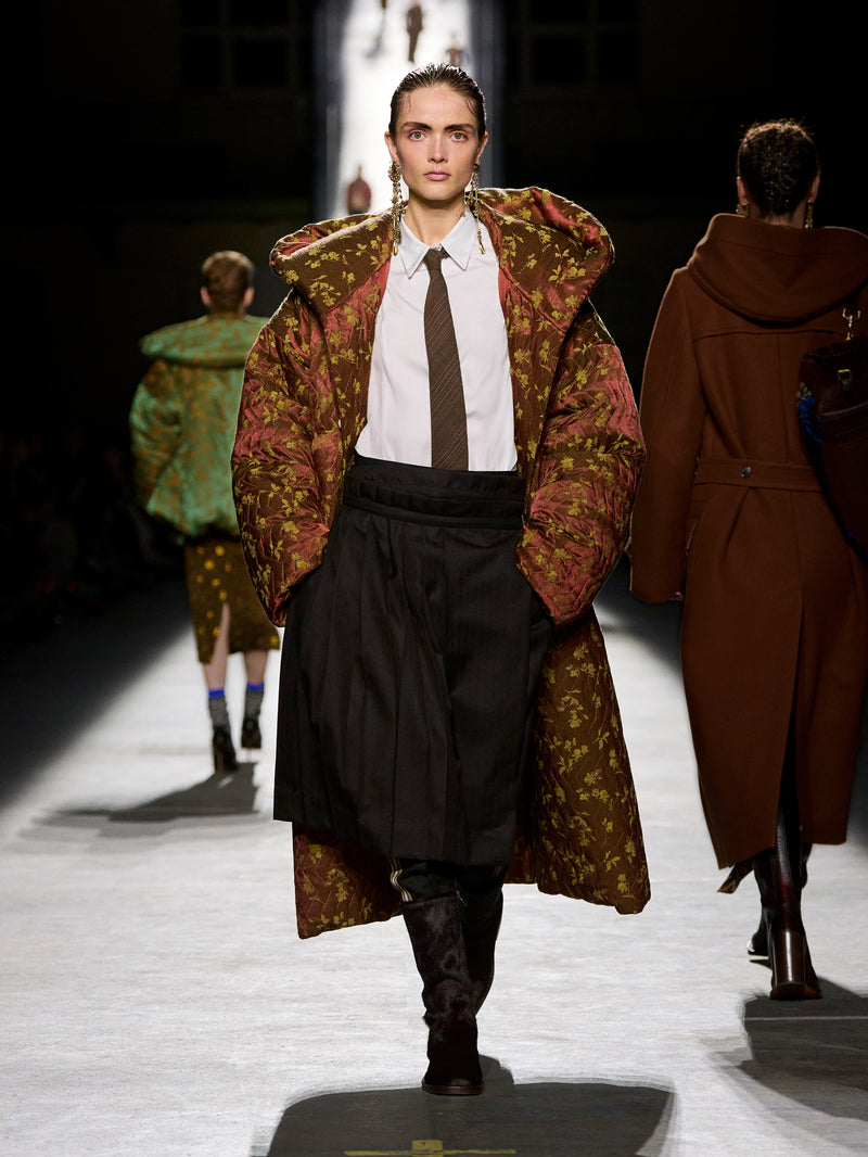 Model walking on a runway wearing a patterned coat and dark pants.
