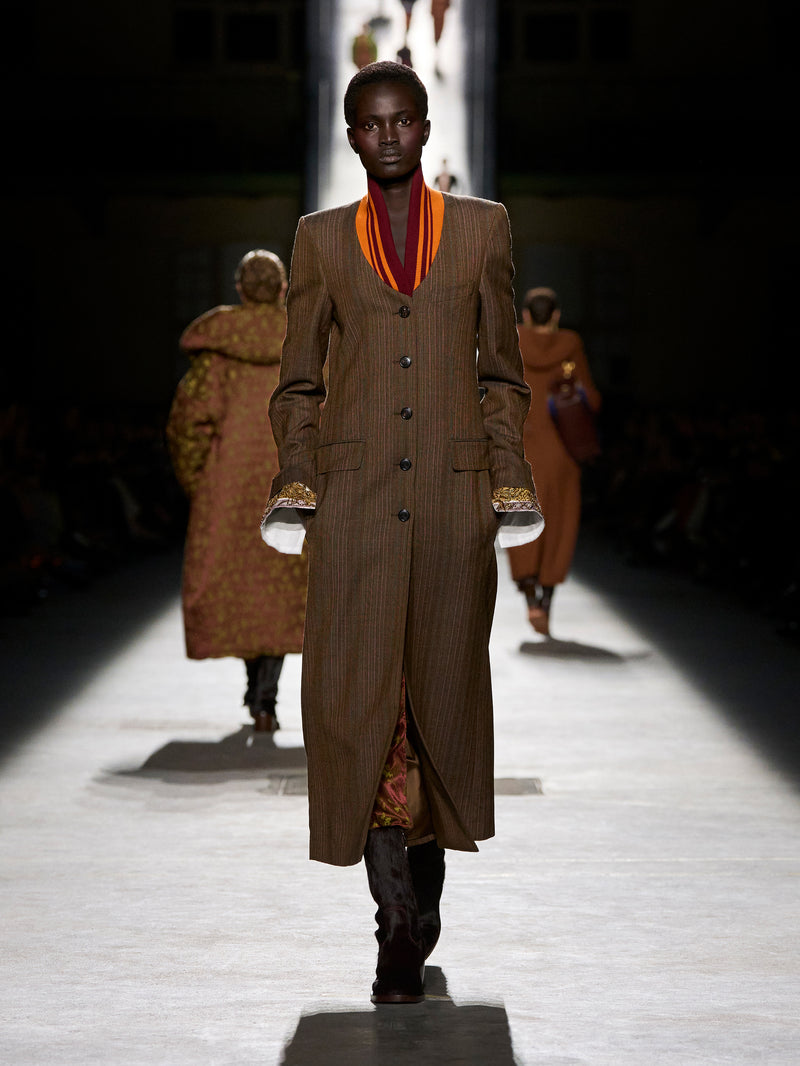 Model walking on a runway wearing a long brown coat with a colorful scarf.