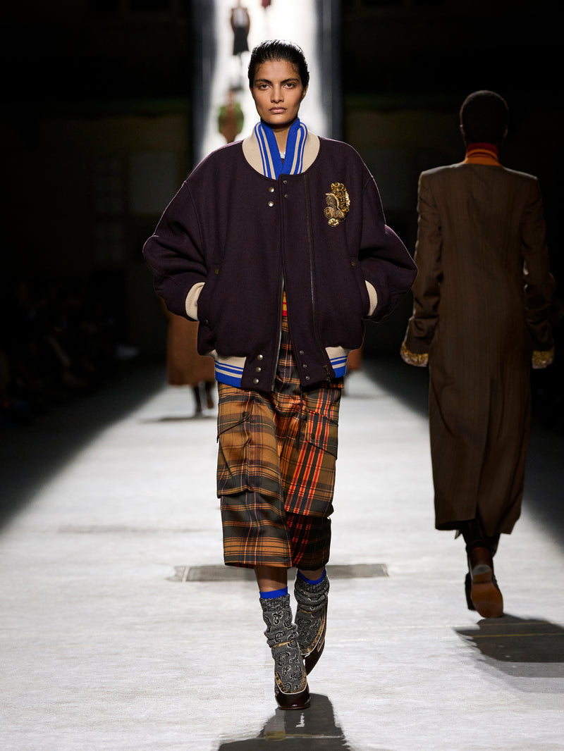 Model walking on a runway wearing a dark jacket with a plaid skirt and patterned socks.