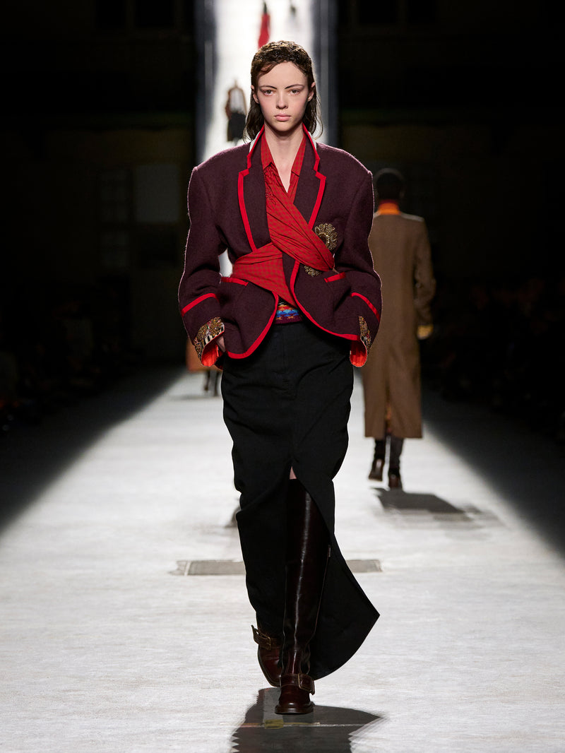 Model walking on a runway wearing a dark outfit with red accents