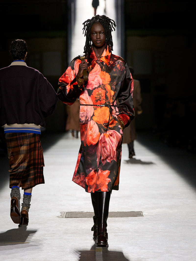 Model walking on a runway wearing a floral dress with a dark background