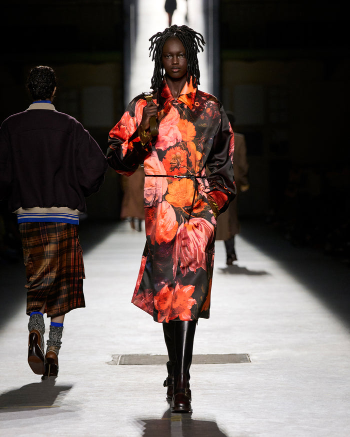 Model walking on a runway wearing a floral dress with a dark background