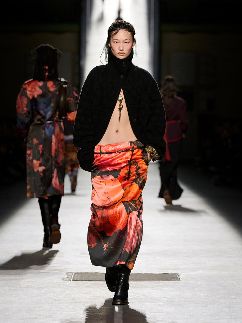 Model walking on a runway wearing a black jacket and colorful skirt.