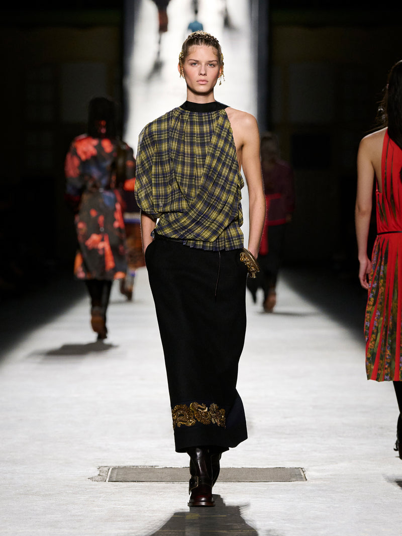 Model walking on a runway wearing a plaid top and black pants with gold accents.