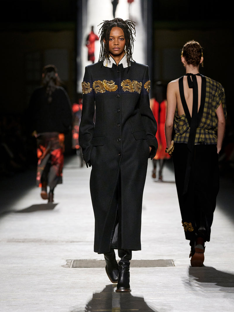 Model walking on a runway wearing a black outfit with gold embroidery.