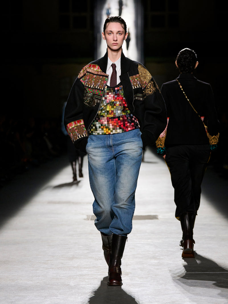 Model walking on a runway wearing a colorful jacket, patterned top, and jeans.