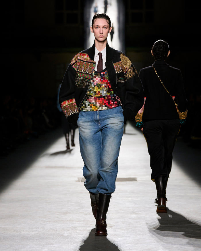 Model walking on a runway wearing a colorful jacket, patterned top, and jeans.