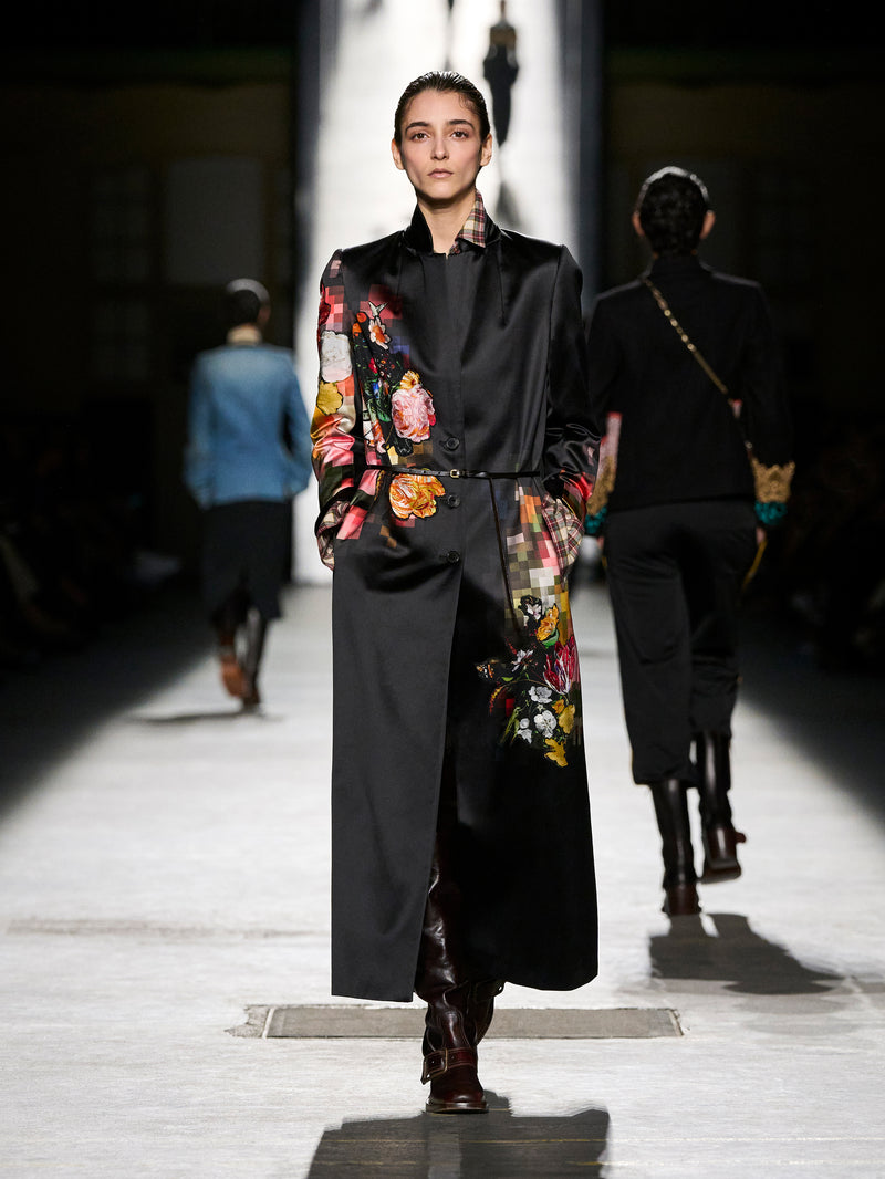 Model walking on a runway wearing a black outfit with floral patterns.