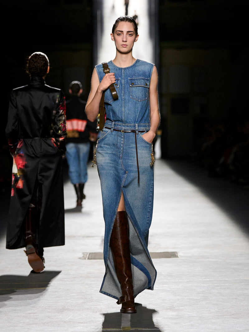 Model walking on a runway wearing a long denim dress with a high slit.