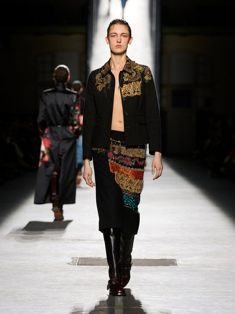 Model walking on a runway wearing a black outfit with colorful patterns.