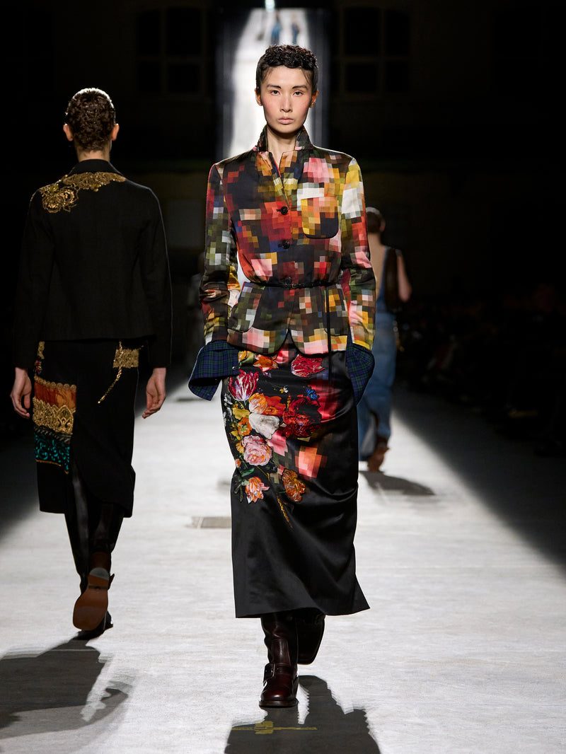 Model walking on a runway wearing a colorful floral dress with a dark background.